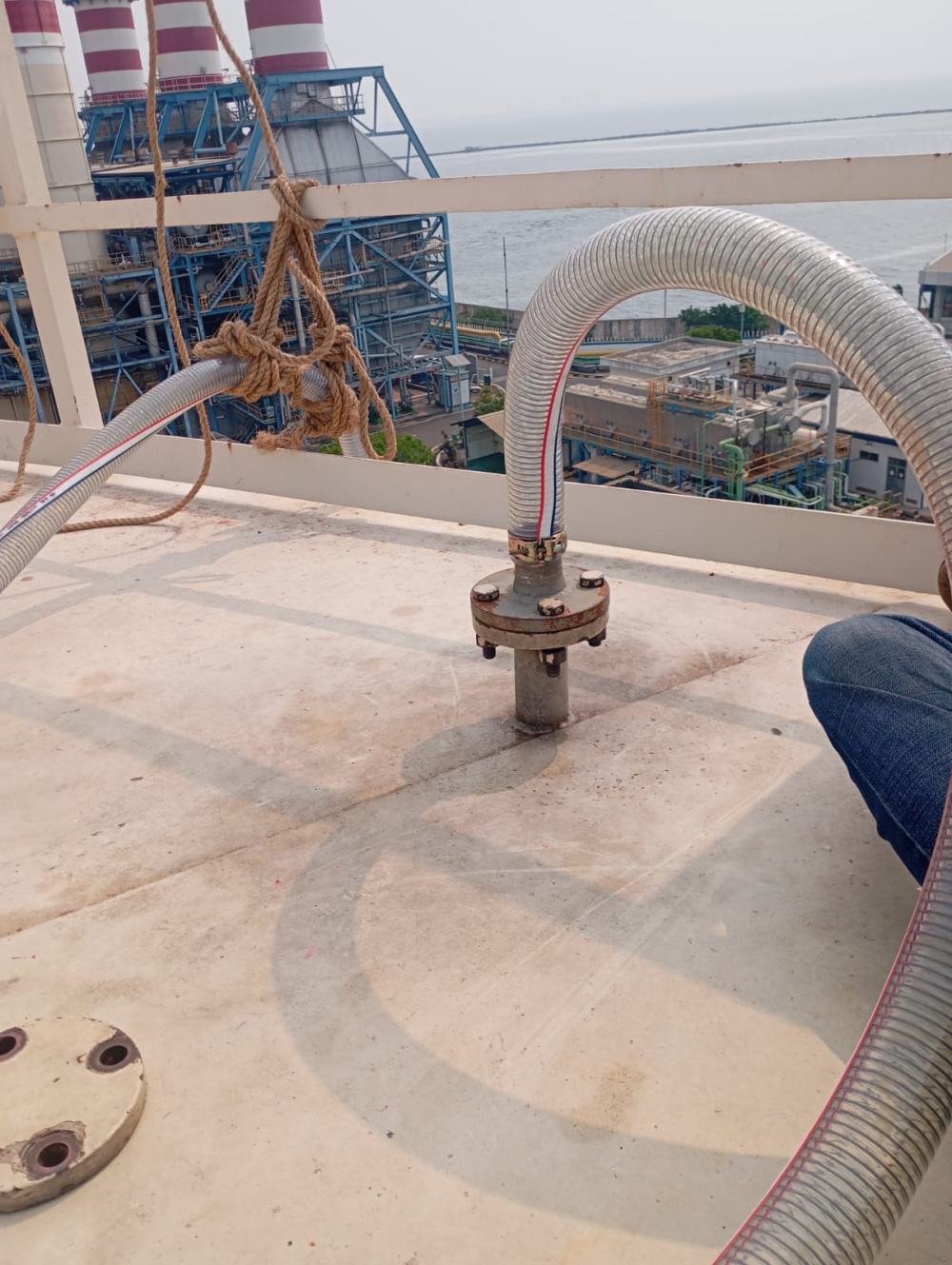 Sambungan hose transfer HSD di atas ground tank PLTU Tanjung Priok dengan latar cerobong pembangkit dan laut